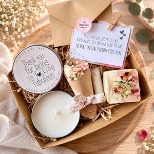 May include: A gift box containing a round candle in a metal tin with the text Thank you for being so f*cking fabulous, a small bouquet of dried flowers wrapped in brown paper, a lip balm, and a bar of soap with dried flower petals on top.