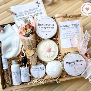 May include: A gift box containing items for a new mother, including a candle, body butter, foot soak, hand cream, body oil, dried flowers, socks, and a card. The items are arranged on shredded paper. The box is tied with a ribbon.