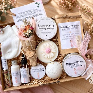 May include: A gift box containing a pair of white baby socks, a dried flower bouquet, a candle labelled Beautiful Mummy to be, a bag of foot soak, a daisy-shaped candle, a bath bomb, hand cream, belly oil, and a tin of stretch mark balm.