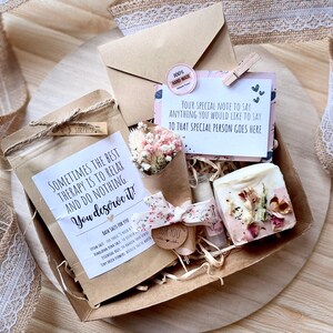 May include: A brown gift box filled with a variety of self-care items, including bath salts, a bar of soap, dried flowers, and a handwritten note. The box is decorated with a brown ribbon and a tag that says "You deserve it!"