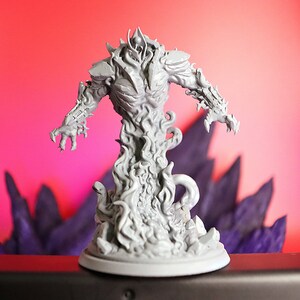 Op de afbeelding: Een grijs, gedetailleerd miniatuur van een vuurwezen met een drakenkop en een basis. Het wezen is geposeerd met zijn armen uitgestrekt, alsof het in een verdedigende houding staat. Het miniatuur is geplaatst tegen een rode en paarse achtergrond.