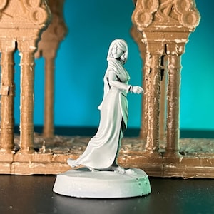 Puede incluir: Una figura en miniatura pintada de blanco de una mujer que lleva una larga túnica fluida y una capa con capucha. La figura está de pie sobre una base redonda y está posada con una mano levantada.