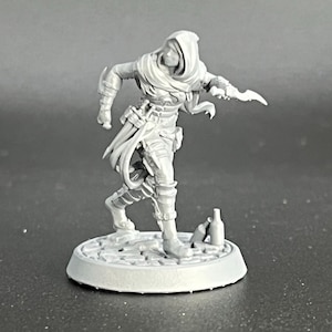 Może przedstawiać: Szara plastikowa figurka miniaturowa osoby w kapturze trzymającej sztylet, stojąca na okrągłej podstawie z brukowanym wzorem. Figurka ma na sobie płaszcz z kapturem i pasek z sakiewką. Na podstawie leżą dwie butelki.