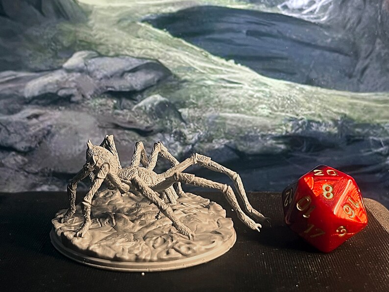 May include: A grey, detailed miniature of a spider with eight legs, standing on a round base. The spider is posed in a crawling position with its front legs raised. A red 20-sided die with gold numbers is in the background.