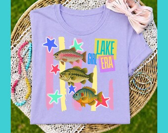 May include: A light purple t-shirt featuring three colorful fish illustrations and the text "LAKE GIRL ERA." The design includes stars and vertical stripes in various colors. The shirt is displayed on a woven surface with accessories.