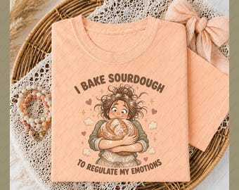 May include: Peach-colored t-shirt with the text "I bake sourdough to regulate my emotions." The graphic features a cartoon of a person hugging a loaf of bread, surrounded by hearts and stars. The shirt is folded on a wicker tray.