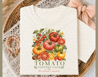 May include: Cream-colored t-shirt featuring a watercolor illustration of various tomatoes, including red, orange, and green varieties, with green leaves and yellow flowers. The text "TOMATO TASTING CLUB" and "heirloom season" are printed below the illustration.