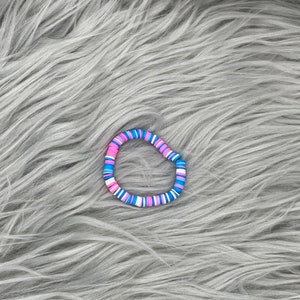 May include: A colorful beaded bracelet with pink, blue, and white beads. The bracelet is made of round, flat beads that are strung together in a repeating pattern.