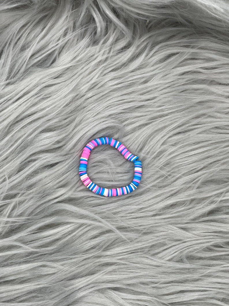 May include: A colorful beaded bracelet with pink, blue, and white beads. The bracelet is made of round, flat beads that are strung together in a repeating pattern.