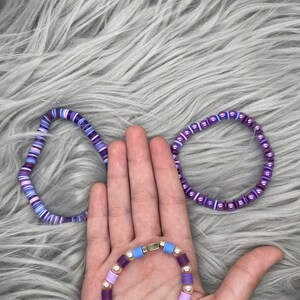 May include: Three purple and lavender beaded bracelets. The bracelets are made with different sized beads and have a variety of colors, including purple, lavender, and white.