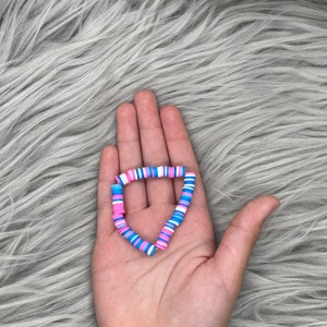 May include: A bracelet made of colorful, round, flat beads. The beads are pink, blue, and white. The bracelet is in the shape of a heart.
