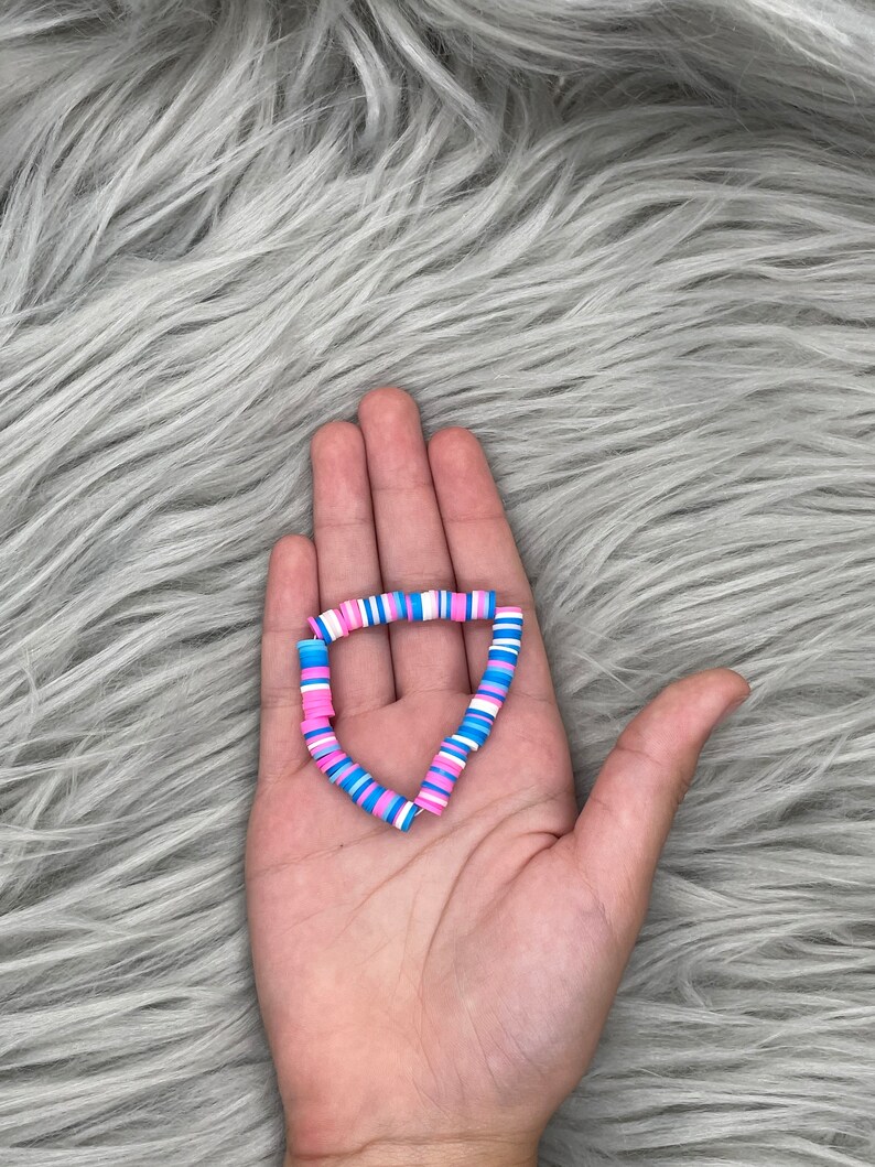 May include: A bracelet made of colorful, round, flat beads. The beads are pink, blue, and white. The bracelet is in the shape of a heart.