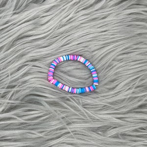 May include: A colorful bracelet made of small, round beads in shades of pink, blue, and white. The bracelet is lying on a gray, fuzzy surface.