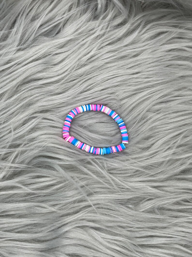 May include: A colorful bracelet made of small, round beads in shades of pink, blue, and white. The bracelet is lying on a gray, fuzzy surface.