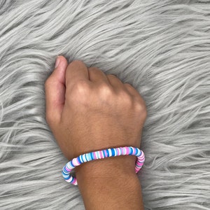 May include: A colorful beaded bracelet with pink, blue, and white beads. The bracelet is made of round, flat beads that are stacked on top of each other.