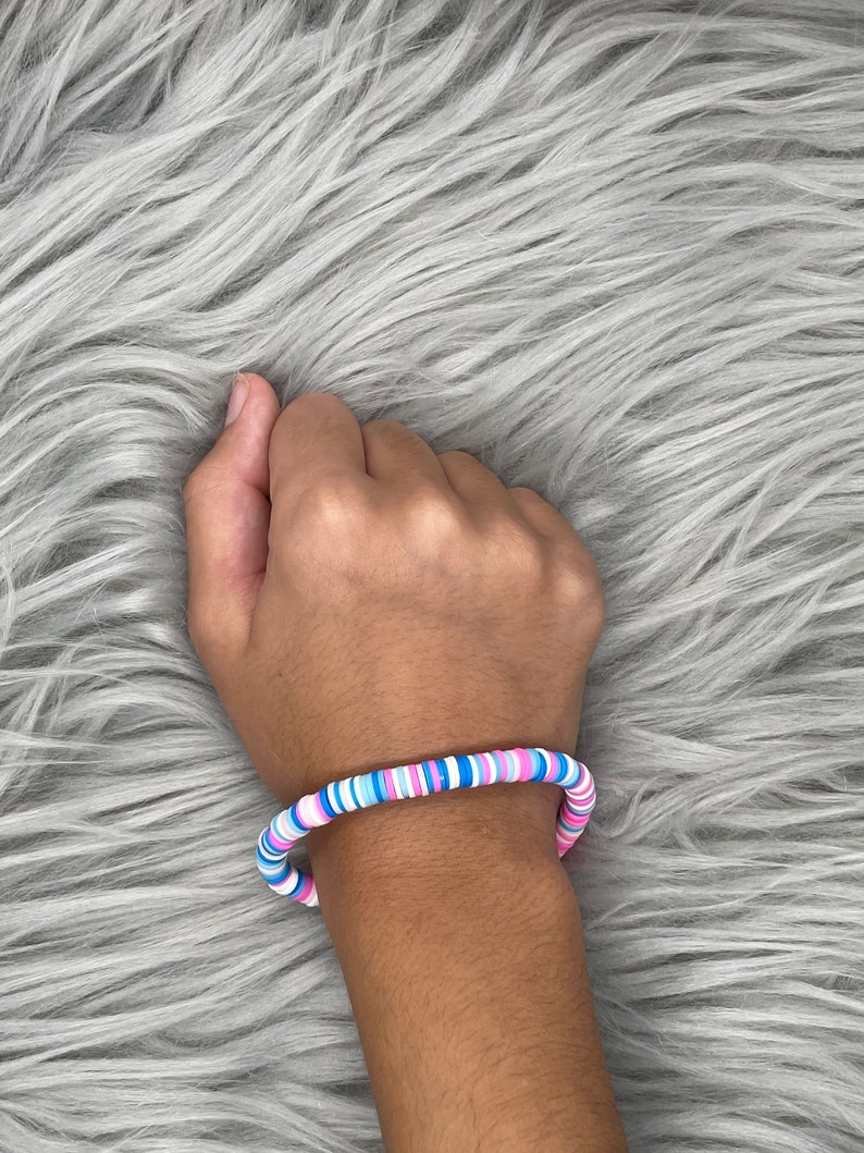 May include: A colorful beaded bracelet with pink, blue, and white beads. The bracelet is made of round, flat beads that are stacked on top of each other.