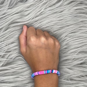 May include: A bracelet made of colorful, round beads. The beads are pink, blue, and white. The bracelet is on a person's wrist.