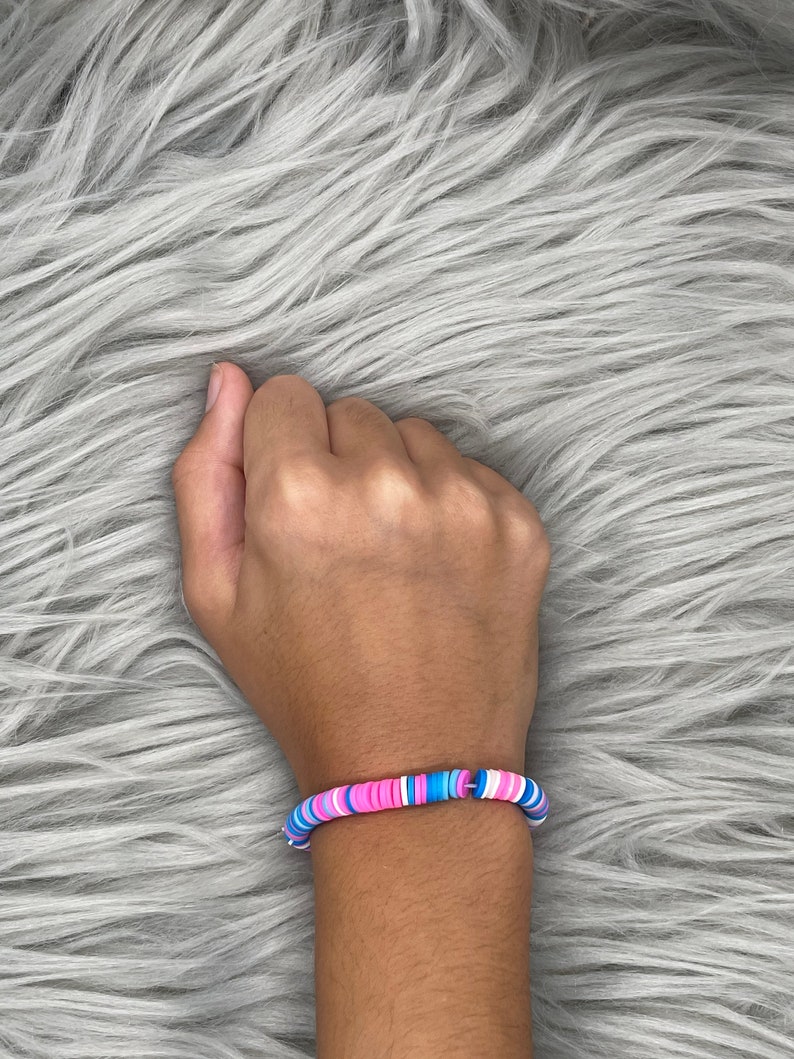 May include: A bracelet made of colorful, round beads. The beads are pink, blue, and white. The bracelet is on a person's wrist.