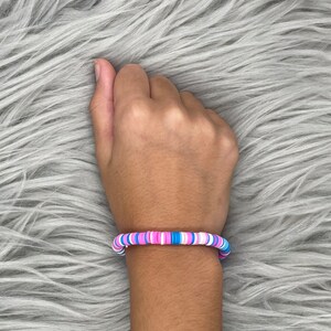 May include: A colorful bracelet made of small, round beads in shades of pink, blue, and white. The bracelet is worn on a person's wrist.