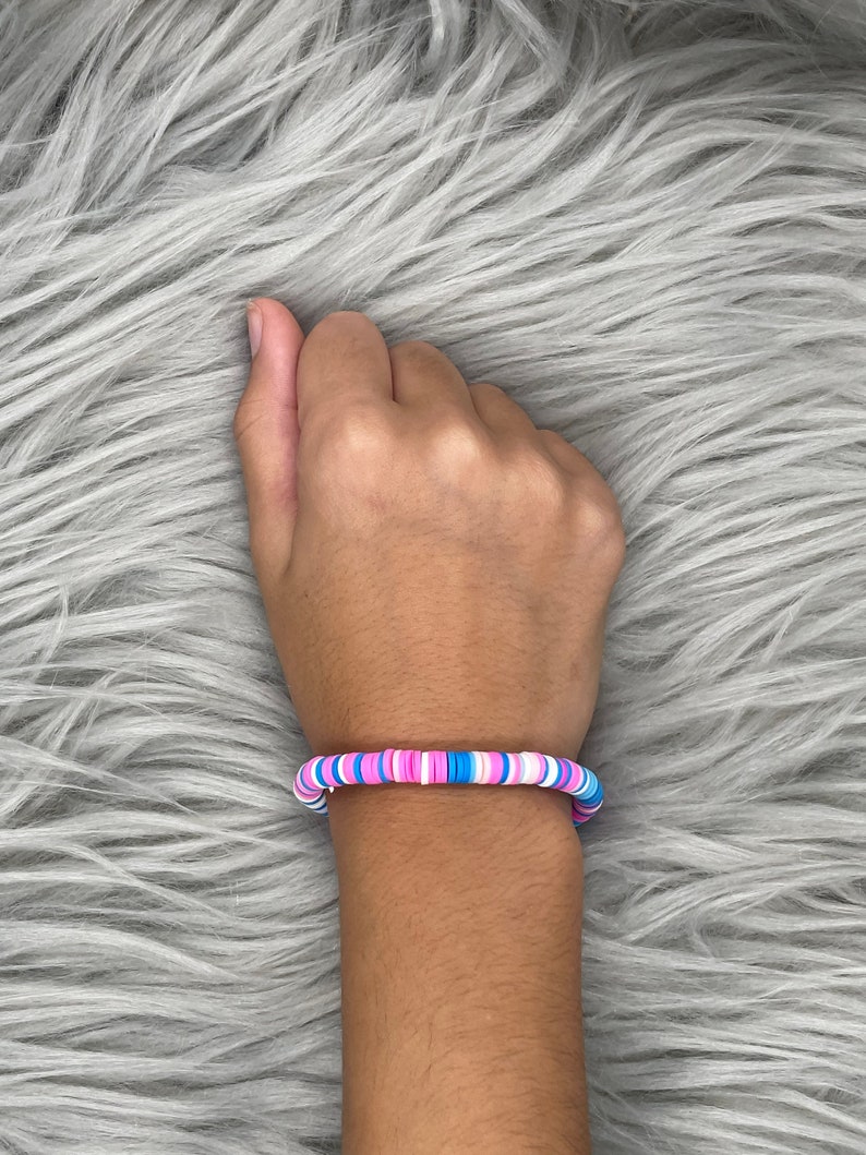 May include: A colorful bracelet made of small, round beads in shades of pink, blue, and white. The bracelet is worn on a person's wrist.