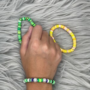 May include: Two bracelets made of colorful round beads. One bracelet is green, white, and blue with two white pearl accents. The other bracelet is yellow, orange, and white.