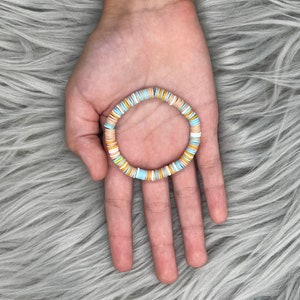 May include: A colorful beaded bracelet with light blue, white, and yellow beads. The bracelet is made of round, flat beads that are strung together to form a circle.