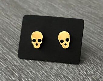 May include: A pair of gold skull stud earrings on a black background.