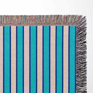 K&ouml;nnte beinhalten: Eine gewebte Wolldecke mit einem gestreiften Muster in Beige, Blau und T&uuml;rkis. Die Decke hat an allen Seiten Fransen.