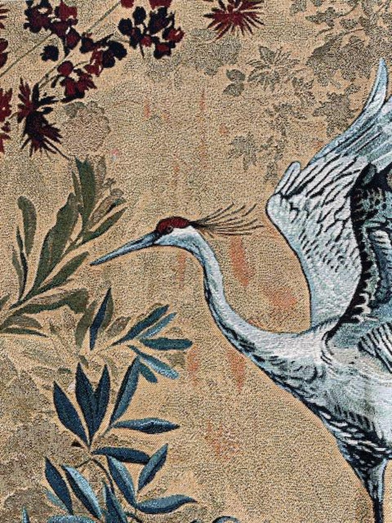 May include: Close-up of a tapestry featuring a crane with outstretched wings, set against a backdrop of floral and foliage patterns. The crane is primarily blue and white, with a red crown. The background is a beige color.