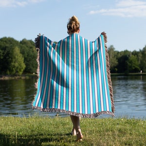 K&ouml;nnte beinhalten: Eine Person steht in einem grasbewachsenen Bereich an einem See und h&auml;lt eine blaue und wei&szlig;e gestreifte Wolldecke mit Fransen.