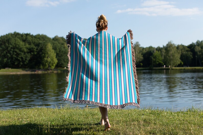 K&ouml;nnte beinhalten: Eine Person steht in einem grasbewachsenen Bereich an einem See und h&auml;lt eine blaue und wei&szlig;e gestreifte Wolldecke mit Fransen.