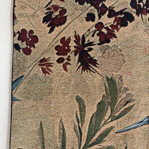 May include: Close-up of a tapestry with a beige background and a floral pattern. The design features burgundy flowers and green leaves, with accents of blue. The tapestry has a black border.