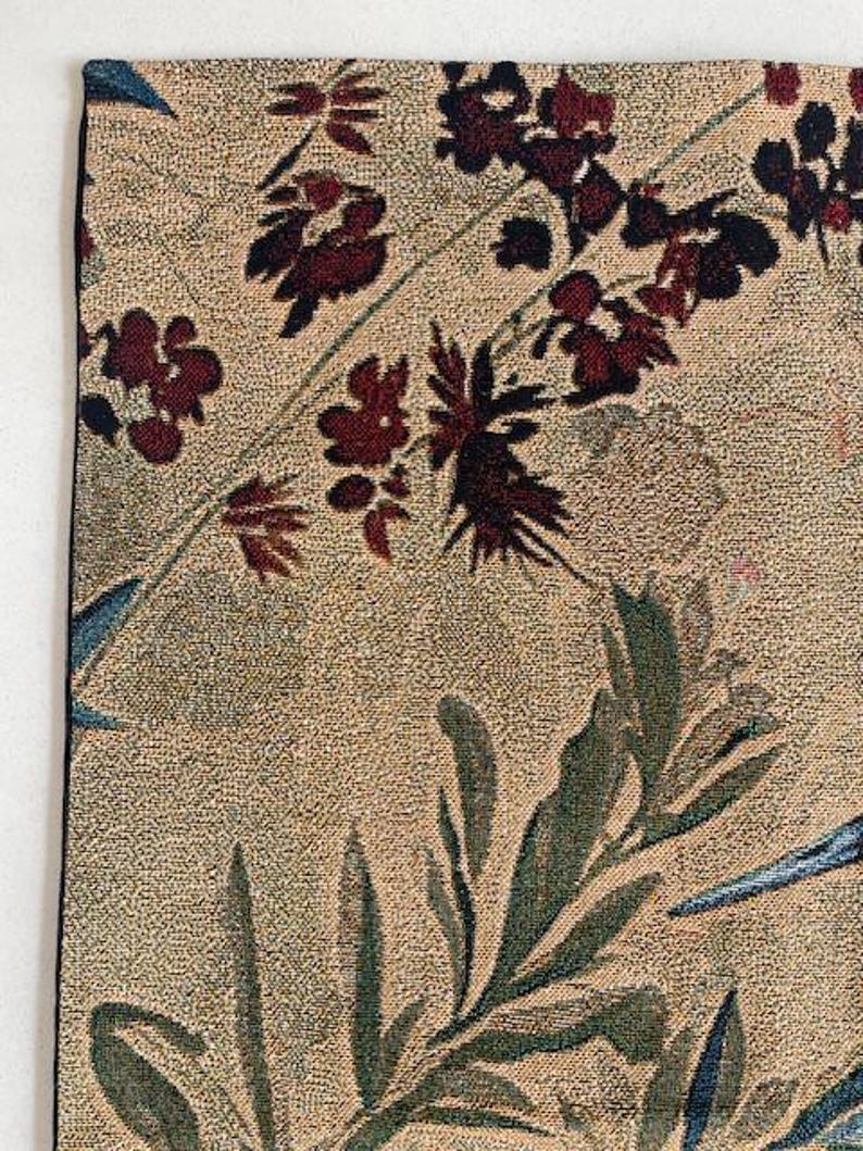 May include: Close-up of a tapestry with a beige background and a floral pattern. The design features burgundy flowers and green leaves, with accents of blue. The tapestry has a black border.