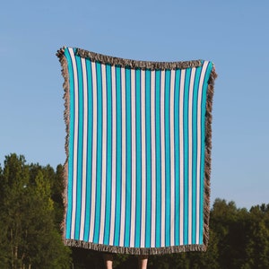 K&ouml;nnte beinhalten: Eine Person h&auml;lt eine blaue und wei&szlig;e gestreifte Wolldecke mit Fransen hoch. Die Decke wird vor dem K&ouml;rper der Person gehalten und verdeckt Gesicht und Oberk&ouml;rper. Die Person steht auf einer grasbewachsenen Wiese mit B&auml;umen im Hintergrund.