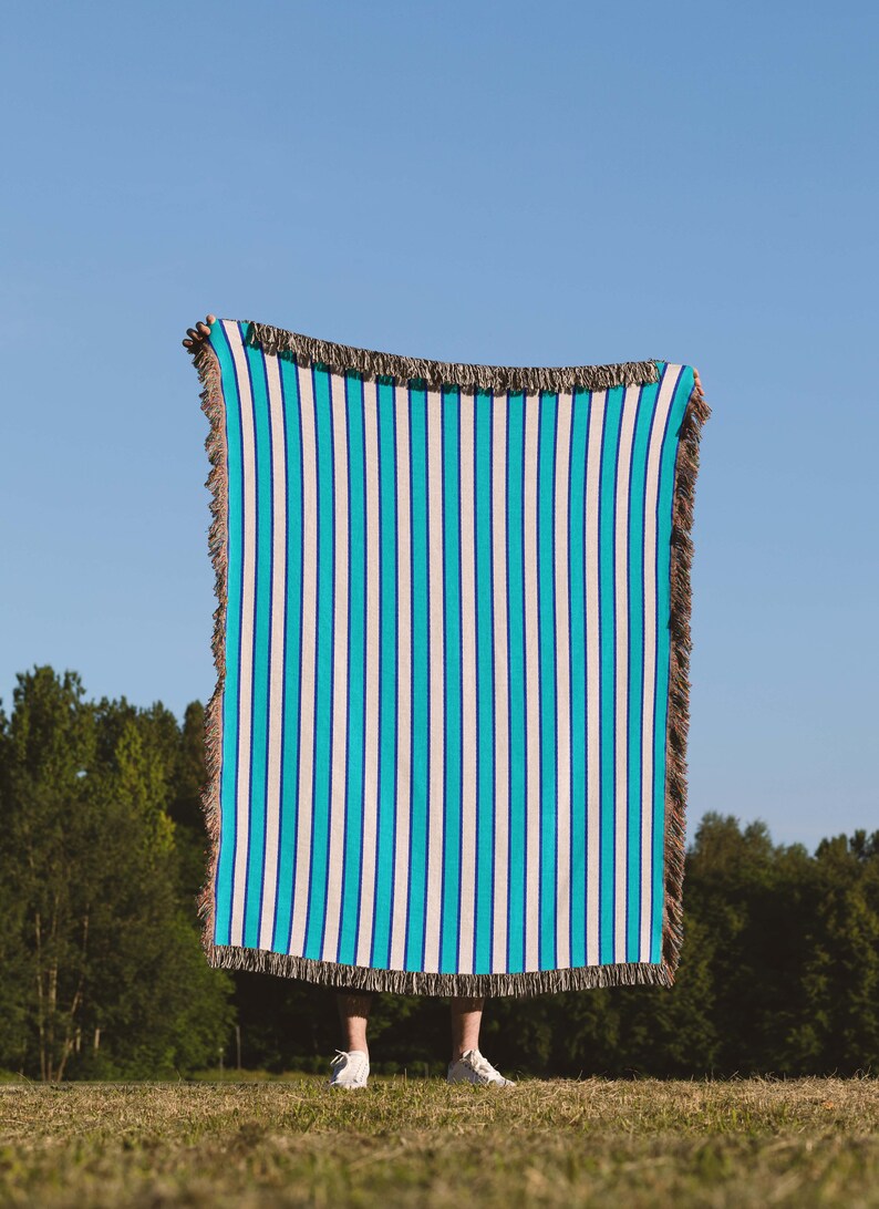 K&ouml;nnte beinhalten: Eine Person h&auml;lt eine blaue und wei&szlig;e gestreifte Wolldecke mit Fransen hoch. Die Decke wird vor dem K&ouml;rper der Person gehalten und verdeckt Gesicht und Oberk&ouml;rper. Die Person steht auf einer grasbewachsenen Wiese mit B&auml;umen im Hintergrund.