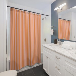K&ouml;nnte beinhalten: Ein Badezimmer mit einem wei&szlig;en Waschtisch und einer Duschvorhang mit orangefarbenen und wei&szlig;en Streifen. Der Duschvorhang ist aus Stoff und hat ein schlichtes Design.