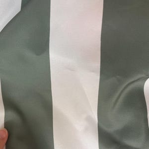 Peut inclure: Gros plan sur un tissu &agrave; rayures verticales. Des rayures larges altern&eacute;es, blanches et vert-gris, cr&eacute;ent un motif simple mais &eacute;l&eacute;gant. Le tissu semble lisse et l&eacute;g&egrave;rement r&eacute;fl&eacute;chissant.