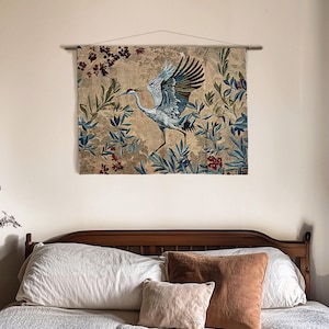 May include: A wall tapestry featuring a crane with outstretched wings, set against a backdrop of foliage in shades of blue, green, and red. The tapestry is mounted on a wooden rod and hangs above a bed with pillows.