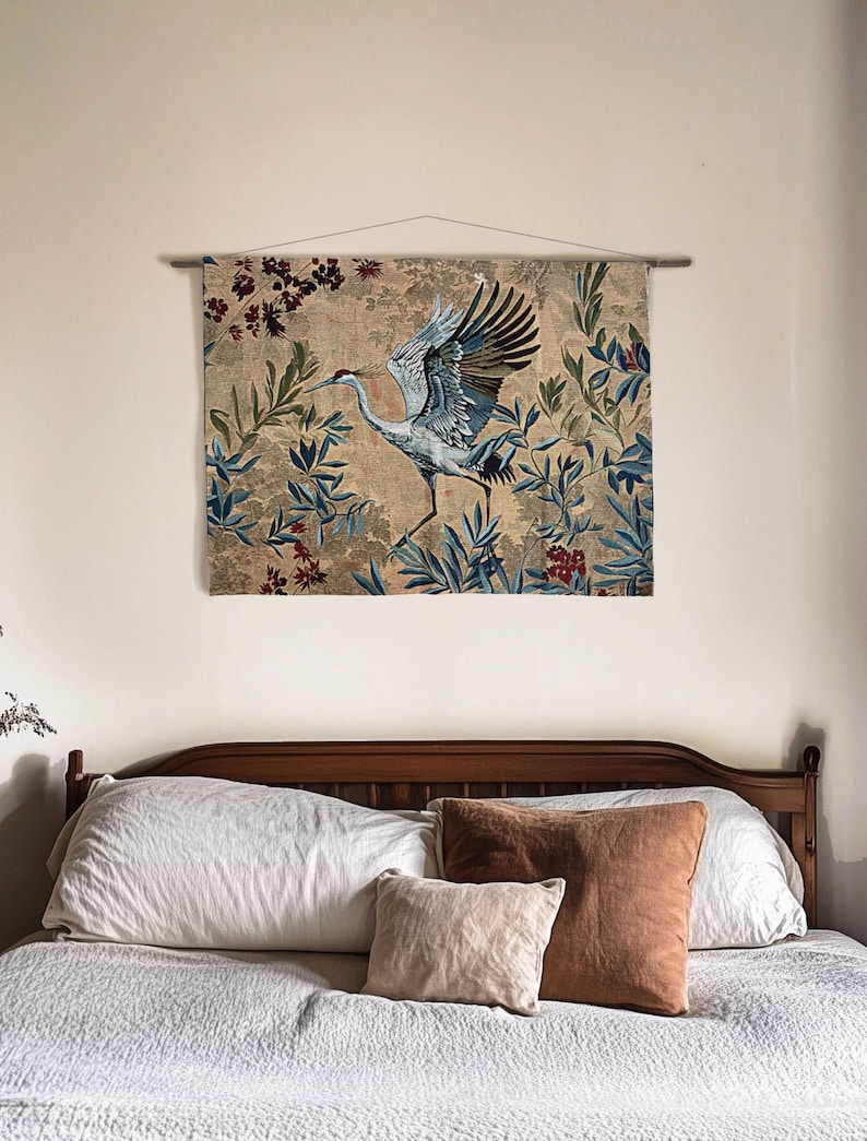 May include: A wall tapestry featuring a crane with outstretched wings, set against a backdrop of foliage in shades of blue, green, and red. The tapestry is mounted on a wooden rod and hangs above a bed with pillows.