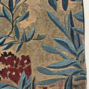May include: Close-up of a woven textile with a floral pattern. The design features teal leaves and burgundy flowers against a tan background. The fabric has a textured appearance, suggesting a tapestry or upholstery material.