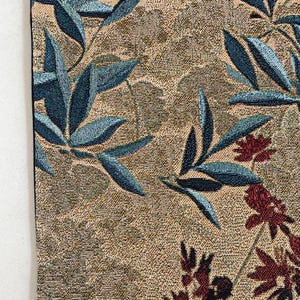 May include: Close-up of a woven textile with a botanical design. The fabric features a tan background with blue-green leaves and burgundy floral accents. The texture of the fabric is visible, adding depth to the design.