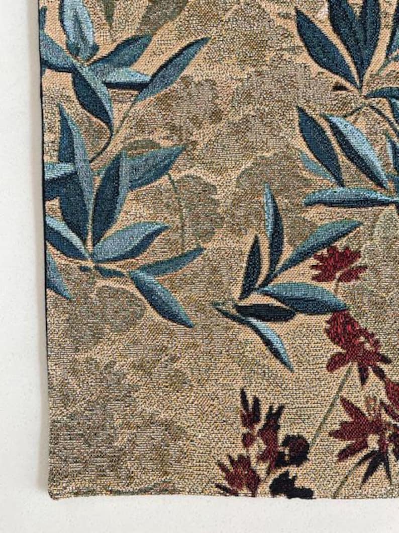 May include: Close-up of a woven textile with a botanical design. The fabric features a tan background with blue-green leaves and burgundy floral accents. The texture of the fabric is visible, adding depth to the design.