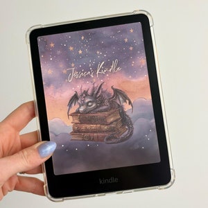 May include: A black and white Kindle e-reader with a clear case. The screen displays a purple and pink watercolor background with gold stars and a small dragon sleeping on a stack of books. The text "Jessica's Kindle" is written in gold cursive on the screen.