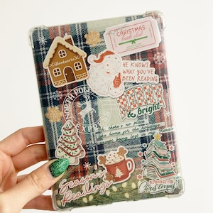 May include: A clear phone case with a plaid pattern and various Christmas-themed stickers. The stickers include a gingerbread house, a Santa Claus, a Christmas tree, a mug of hot chocolate, a stack of books, and the text "Season's Readings" and "All Booked for Christmas".
