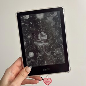Pode incluir: Um leitor de e-books Kindle preto e branco com uma capa transparente. A tela exibe uma imagem em preto e branco de uma cena celestial com o texto "Just One More Chapter". Um amuleto em forma de coração rosa com o texto "Emotional Support Kindle" pendura na parte inferior do leitor de e-books.