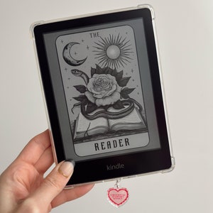 May include: A black and white illustration of a tarot card with a rose, a snake, a moon, and a sun. The card is labeled "The Reader". The card is displayed on a Kindle e-reader with a pink heart charm attached to the bottom.