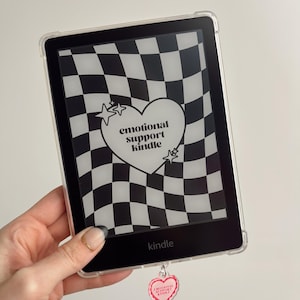 Puede incluir: Un lector de libros electrónicos Kindle negro con una funda protectora transparente. La pantalla muestra un patrón de cuadros en blanco y negro con un corazón que dice "emotional support kindle". Un dije en forma de corazón con el mismo texto cuelga del dispositivo.