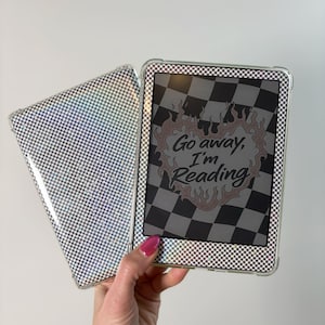 May include: An e-reader with a clear case and a matching cover, both featuring a checkered pattern. The screen displays the text "Go away, I'm Reading" within a flame-like design. The case is held by a hand with pink nail polish.