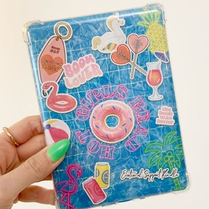 May include: A clear case for an e-reader with a blue and white pool-themed design. The case has several stickers on it, including a pink flamingo, a donut, a pineapple, and a unicorn. The text "Emotional Support Kindle" is on the case.