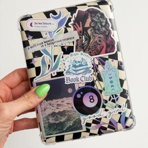 Peut inclure: Une coque de téléphone transparente avec un motif à carreaux noirs et blancs. La coque est décorée de divers autocollants, dont une femme lisant un livre, un crâne avec un livre, une lune et des étoiles, une bougie, une fleur, une photo d'un paysage marin et une boule noire et blanche avec le texte "good girl".